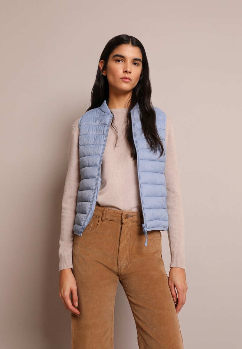Gilet imbottito azzurro chiaro con design a trapunte verticali, chiusura con zip e un maglione in maglia beige a contrasto. Pantaloni in corduroy nei toni della terra.