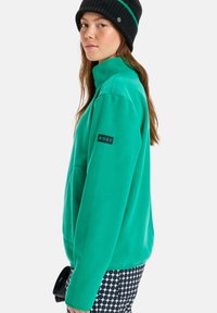 Casaco de fleece verde com colarinho alto e logo da Roxy no braço esquerdo. Combinado com calças estampadas em preto e branco. Gorro preto na cabeça.