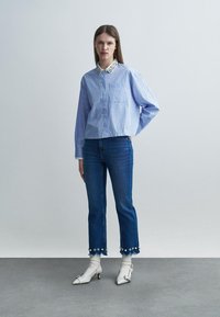 Mujer con una camisa azul a rayas con cuello floral, jeans recortados con adornos de flores, calcetas blancas y zapatos blancos de tacón.