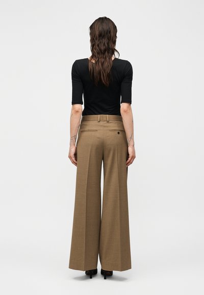 Γυναίκα με μακριά καστανά μαλλιά στέκεται με την πλάτη, φοράει μαύρο τοπ μέχρι τον αγκώνα και καφέ φαρδιά παντελόνια με μαύρες ψηλοτάκουνες.