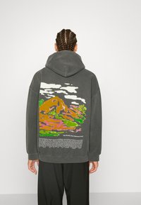 Felpa grigia oversize con stampa di un paesaggio montano colorato sul retro, con dettagli verdi, arancioni e rosa, e testo sotto il design.