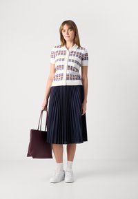 Gilet blanc à motif carreauté, jupe plissée bleu marine, baskets blanches et sac fourre-tout bordeaux. Textures lisses et designs structurés.