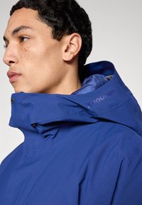 Blå vattentät jacka med hög krage och en åtsittande huva. Har synligt varumärke längs axeln och texturerat tyg.