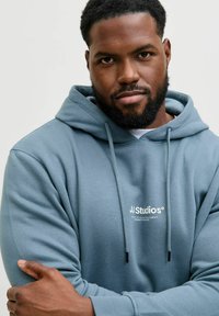 Sweat à capuche bleu clair avec logo blanc, texture douce et cordons réglables. Présente une coupe décontractée et une poche kangourou.