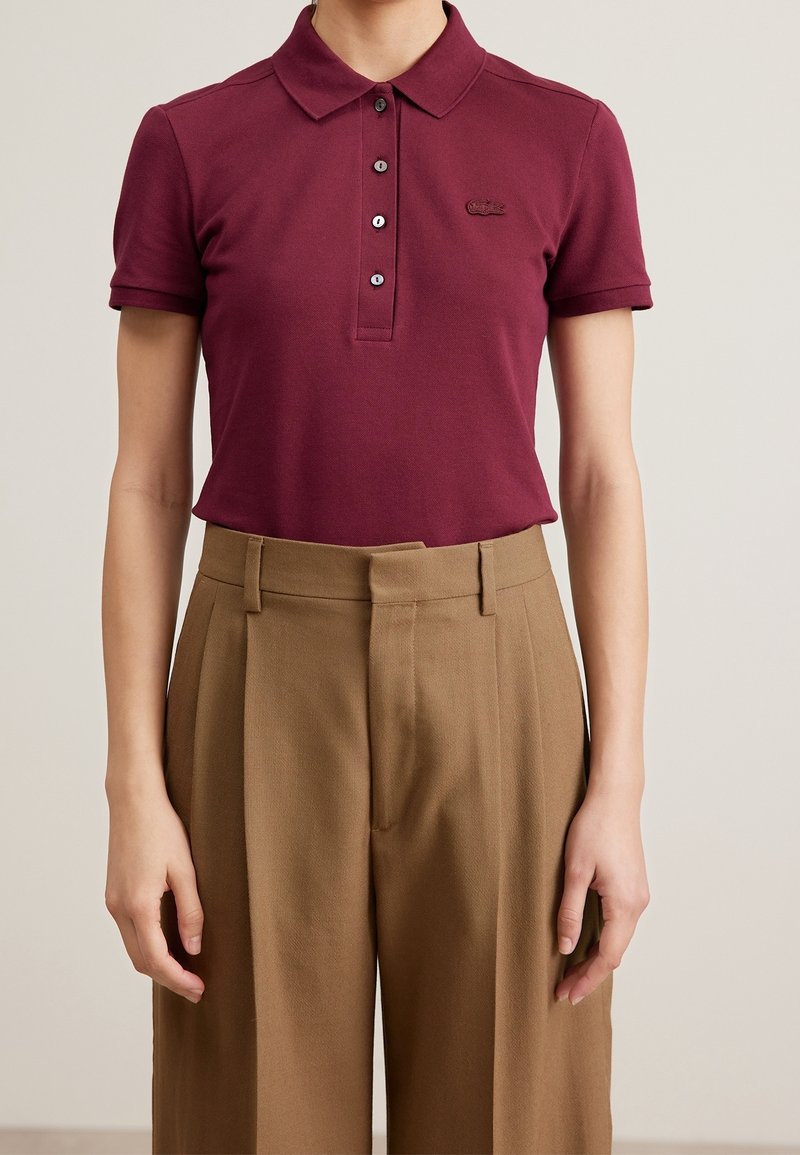 Personne portant un polo bordeaux à manches courtes avec des boutons et un pantalon habillé brun taille haute sur un fond uni.