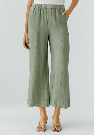 Femme portant un pantalon ample en lin vert olive avec taille élastique et jambes larges plissées, associé à des sandales argentées, debout sur fond blanc.