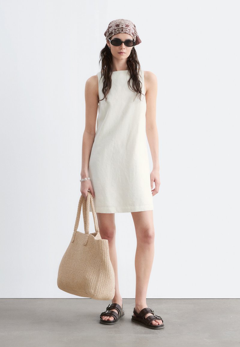 Femme portant une robe blanche sans manches, un foulard à motifs, des lunettes de soleil foncées, portant un grand sac cabas beige tissé et des sandales noires.
