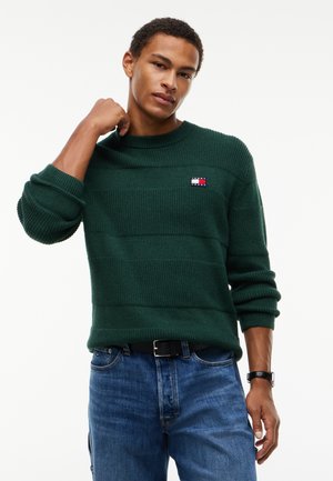 Jonge man draagt een donkergroene gebreide trui met een logo patch en blauwe spijkerbroek, poseert met één hand bij zijn nek en de ander omlaag.