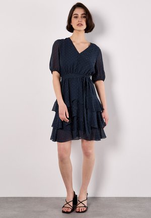 Robe bleu marine à volants fabriquée en tissu léger et semi-transparent avec des pois blancs, dotée d'un décolleté en V et de manches bouffantes. Sandales noires à bretelles.