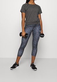 Harmaan lyhythihainen urheilu paita, kuvioidut mustat leggingsit, joissa on tekstiä, ja mustat urheilukengät. Raskaudet pidetään käsissä.