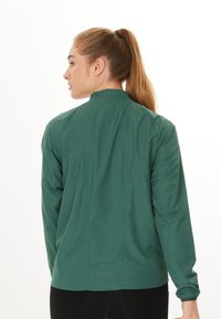 Groene, lichtgewicht jas met hoge kraag, lange mouwen en een subtiele glans. Bevat zijsplitten voor ventilatie en elastische manchetten.