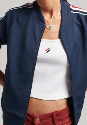 Femme portant une veste zippée bleu marine avec des rayures blanches et rouges sur un top court blanc et un pantalon rouge, avec un collier pendentif rond.