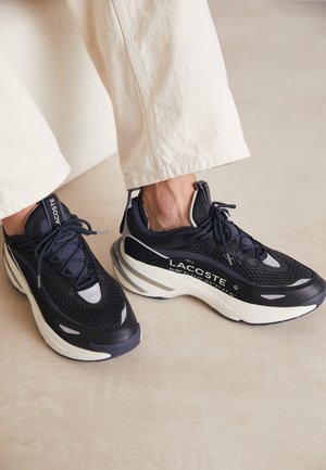 Osoba nosząca czarno-białe sneakersy Lacoste z grubymi podeszwami i białe spodnie, stojąca na jasnobeżowej podłodze.