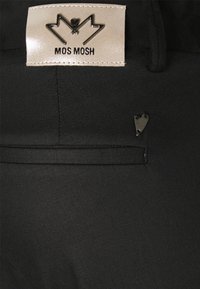 Czarna tkanina o gładkiej fakturze, z metką "MOS MOSH" w srebrnym kolorze oraz małym metalowym serduszkiem na kieszeni.