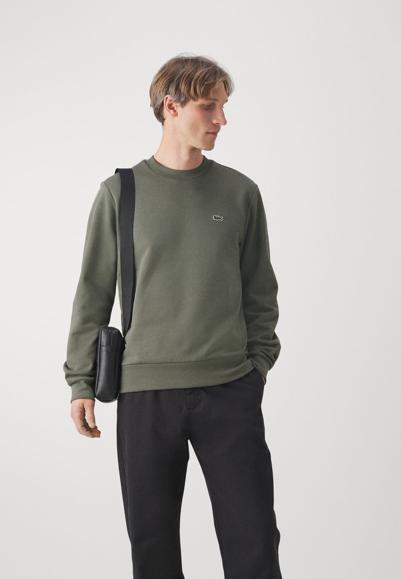 Olijfgroene sweatshirt met lange mouwen en een ronde hals. Bevat een geborduurd logo op de borst en heeft een relaxte pasvorm. Zwarte tas op de schouder.