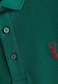 Green polo shirt with a textured knit fabric, featuring three dark buttons and a red deer logo on the chest. Classic collar design.