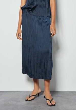 Dark blue pleated midi skirt made of shiny fabric, featuring a loose fit and an elastic waistband, paired with black strappy sandals.