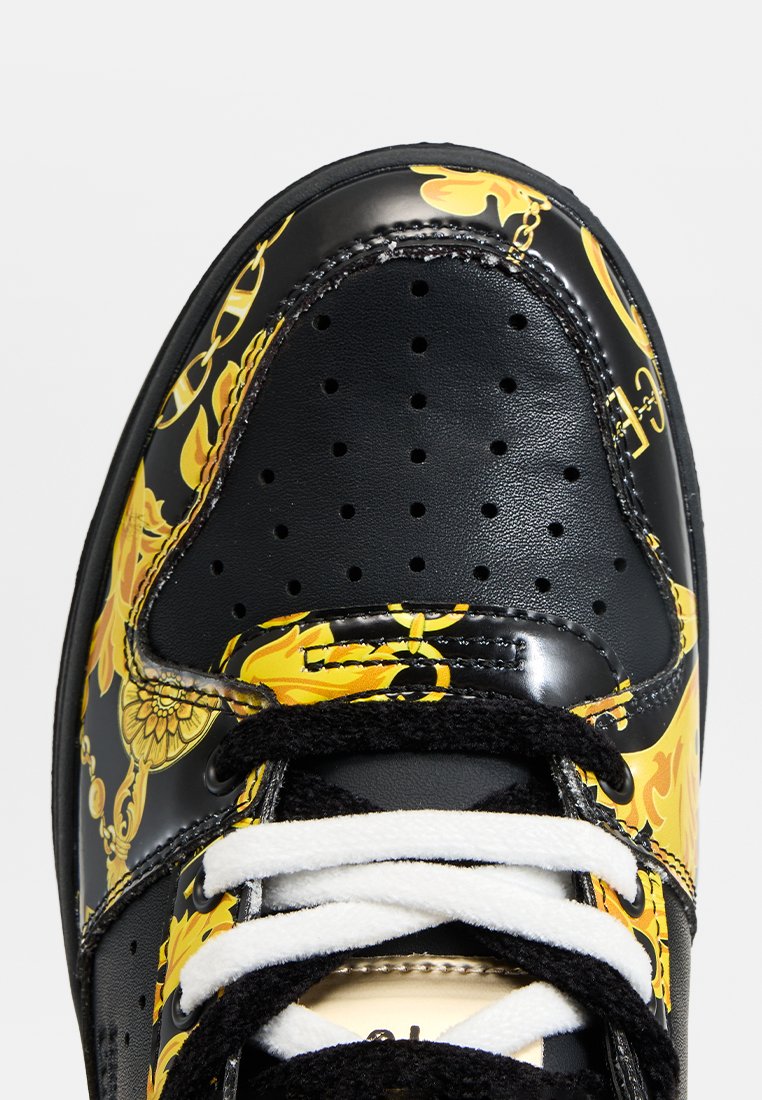 Baskets noires avec un bout en cuir perforé et un motif de chaîne florale jaune, accompagnées de lacets blancs et noirs.