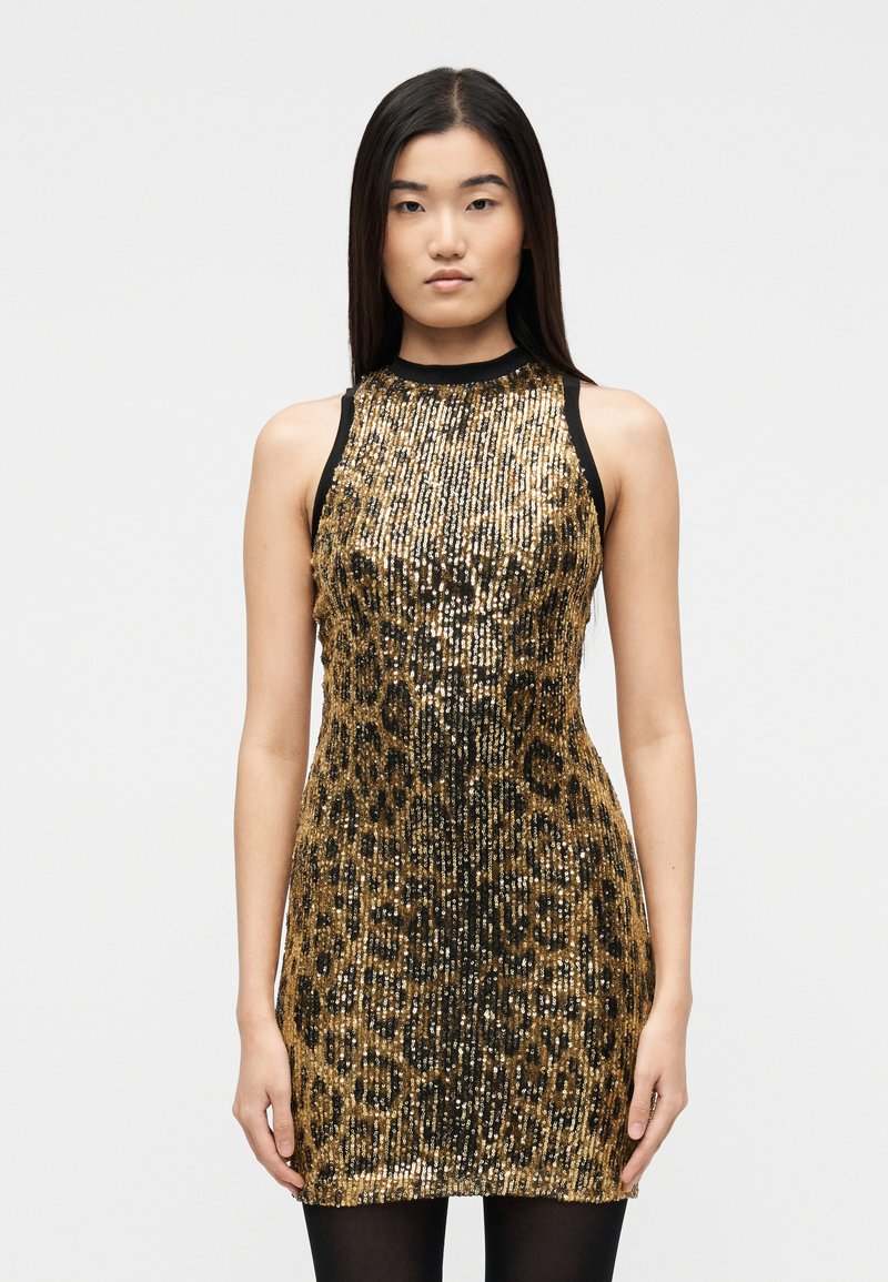 Γυναίκα που φοράει μίνι φόρεμα χωρ ίς μανίκια με χρυσό και μαύρο λεοπάρ τύπωμα από παγιέτες και μαύρες καλσόν, στέκεται μπροστά σε απλό λευκό φόντο.