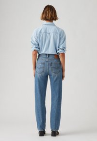 Chemise en denim bleu clair à manches retroussées, associée à un jean droit taille haute. Lavage bleu foncé avec poches arrière visibles et patch en cuir.