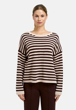Frau mit einem braun-beigen, horizontal gestreiften Langarmpullover und dunkelbraunen Hosen, die nach vorne schaut mit einem neutralen Gesichtsausdruck.
