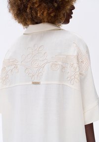 Crème-kleurig shirt, van lichtgewicht stof, met delicate beige borduursels van krullen en vogels op de rug, voorzien van een kraag en korte mouwen.