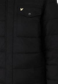 Chaqueta de plumas negra con un bolsillo frontal con solapa, un emblema dorado y paneles cosidos en la tela. Textura suave, acabado mate.