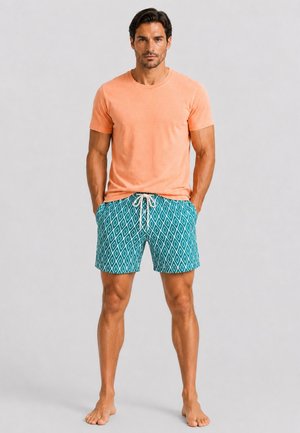 Mann steht barfuß, trägt ein pfirsichfarbenes T-Shirt und blaue gemusterte Shorts mit den Händen in den Taschen vor einem einfarbigen Hintergrund.