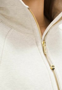Světlý béžový zip-up sweatshirt s vysokým límcem, se zlatým zipem a hladkým povrchem. Zajímavý detail loga na táhle zipu.