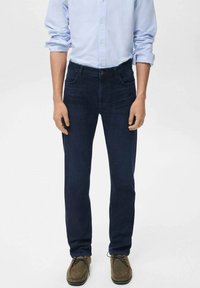 Hombre vestido con jeans oscuros, camisa de botones de color azul claro con las mangas arremangadas y zapatos de cordones casuales marrones, de pie contra un fondo liso.