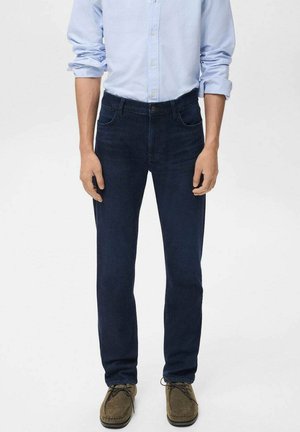 Man in een donkerblauwe jeans, een lichtblauw overhemd met opgerolde mouwen en een paar bruine casual veterschoenen, staande voor een effen achtergrond.