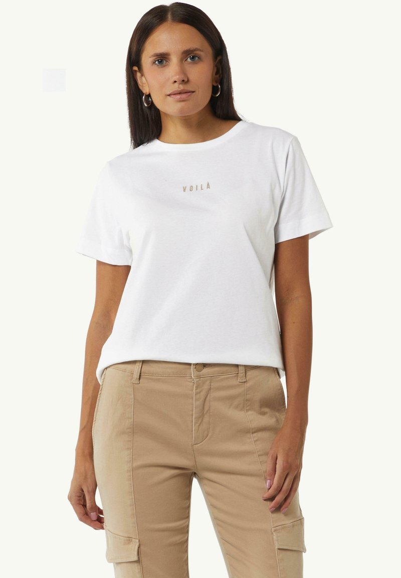 White cotton t-shirt with a round neck and short sleeves, featuring a small gold "VOILA" logo on the chest, paired with beige cargo pants.