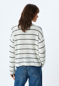 Maglione lavorato a maglia grossa in off-white con strisce navy, caratterizzato da una vestibilità ampia e una texture a coste, abbinato a jeans blu a vita alta.
