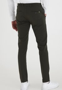 Mann in dunkelgrünen Slim-Fit-Hosen mit Gesäßtaschen, kombiniert mit weißen Turnschuhen und einem weißen Langarmshirt, stehend.