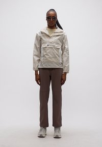 Γυναίκα που φοράει μπεζ αδιάβροχο Columbia, καφέ παντελόνι, γκρι αθλητικά παπούτσια και σκουρόχρωμα γυαλιά ηλίου, στέκεται μπροστά σε απλό γκρι φόντο.