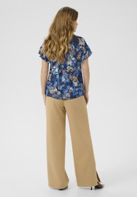 Blomstret blå bluse med korte ærmer, parret med beige brede bukser. Blusen har et levende mønster og en afslappet pasform.