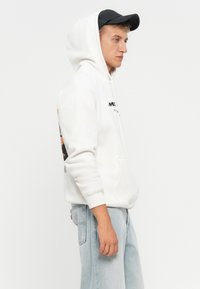 En ung mand iført en hvid hoodie med grafisk print og lyseblå jeans, poserer sidelæns mod en simpel hvid baggrund.