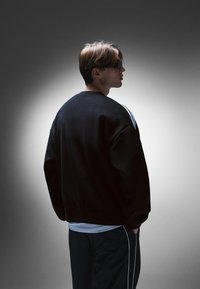 Fekete pulóver kerek nyakkivágással és ejtett vállakkal, fehér csíkokkal az ujjakon. Sötét nadrággal párosítva. Sima textúra.