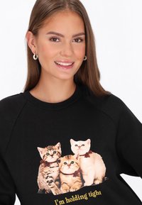 Czarna bluza z nadrukiem trzech kotów, o miękkiej fakturze i okrągłym dekolcie. Pod nadrukiem znajduje się napis "Trzymam mocno."