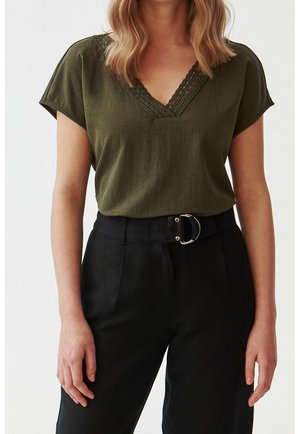 Vrouw die een olijfgroen T-shirt met korte mouwen, een gevlochten V-hals en een zwarte broek met hoge taille draagt, afgewerkt met een zilveren D-ring riem.