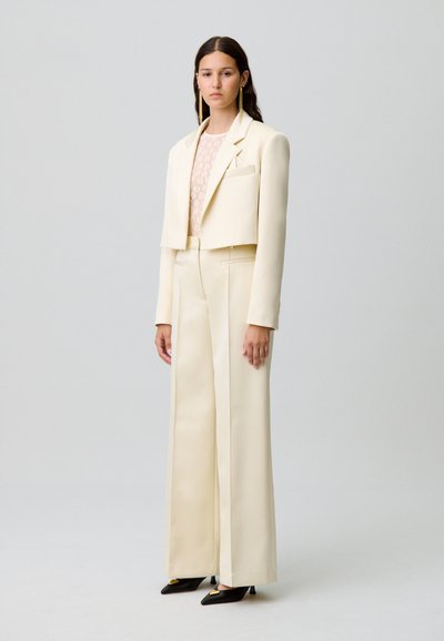 Blazer crème coupé sur un top en dentelle transparente, associé à un pantalon crème à jambes larges et des escarpins noirs à bout pointu avec des accents dorés.