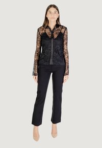 Top in pizzo nero a maniche lunghe con colletto in satin e chiusura a bottoni, abbinato a pantaloni neri aderenti e décolleté nudi.