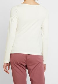 Pitkähihainen, vaalean kermaisen värinen paita, joka on valmistettu sileästä kankaasta. Ominaisuuksia ovat leveä, pyöreä kaula-aukko ja istuva siluetti.