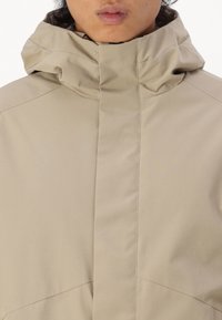 Beige zipjack met hoge kraag, gemaakt van een gladde stof. Heeft strakke lijnen en een minimalistisch ontwerp zonder zichtbare patronen.