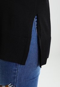 Schwarzer Strickpullover mit gerippten Säumen und einem seitlichen Schlitzdesign. Kombiniert mit blauen Jeans aus Denim, die eine bestickte Akzentuierung zeigen.
