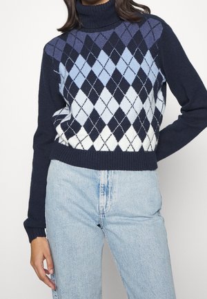 Femme portant un pull à col roulé bleu marine avec un motif losanges bleu et blanc, associé à un jean taille haute bleu clair, debout avec un bras plié.