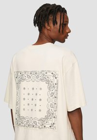 Crème katoenen t-shirt met korte mouwen, met een zwart bandana-print op de achterkant. Losse fit en zachte textuur.