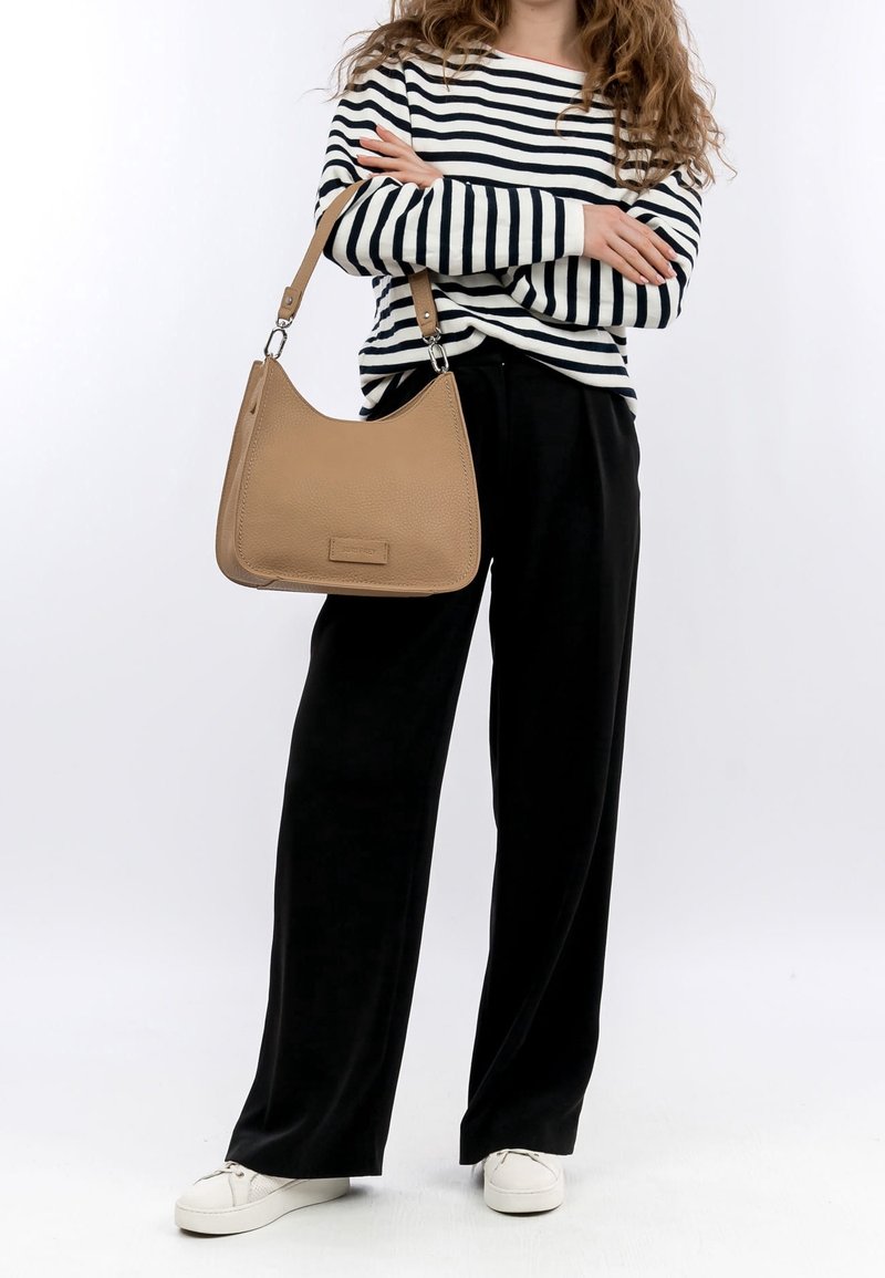 Tan leather shoulder bag with a slightly textured surface, semi-circular shape, and adjustable strap, paired with a striped top and black pants.