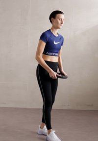 Top corto Nike Pro en azul oscuro, leggings negros con raya blanca en el costado y zapatillas deportivas blancas; sosteniendo una placa de peso circular.