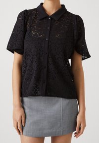 Svart spetsblus med krage, korta puffärmar och knäppning. Bärs med en grå figurnära kjol. Detaljerat blommönster.